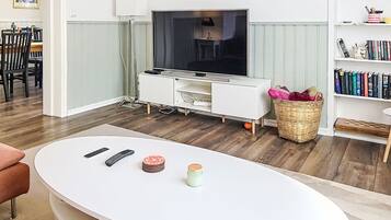 A living room with a TV, white entertainment unit, wooden flooring, a dining area, and a bookshelf.