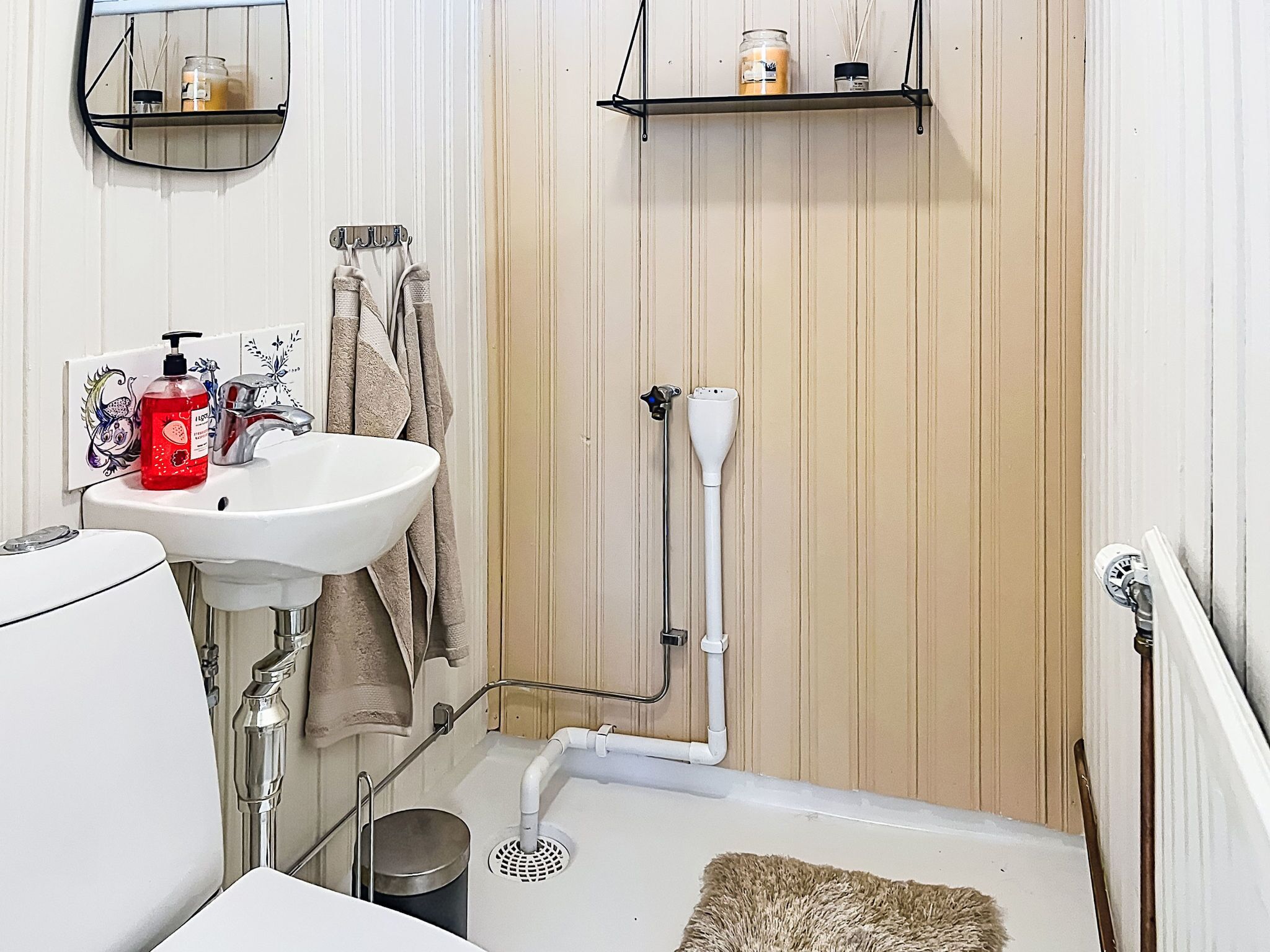Et badeværelse med en hvid vask, et spejl, et håndklædetørrestativ, en bruser og en lille hylde med toiletartikler.