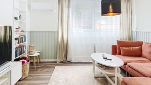 Ein Wohnzimmer mit einer Couch, einem Couchtisch, einem Fernseher, einem Bücherregal, einem Korbwägelchen und einem Fenster mit Vorhängen.