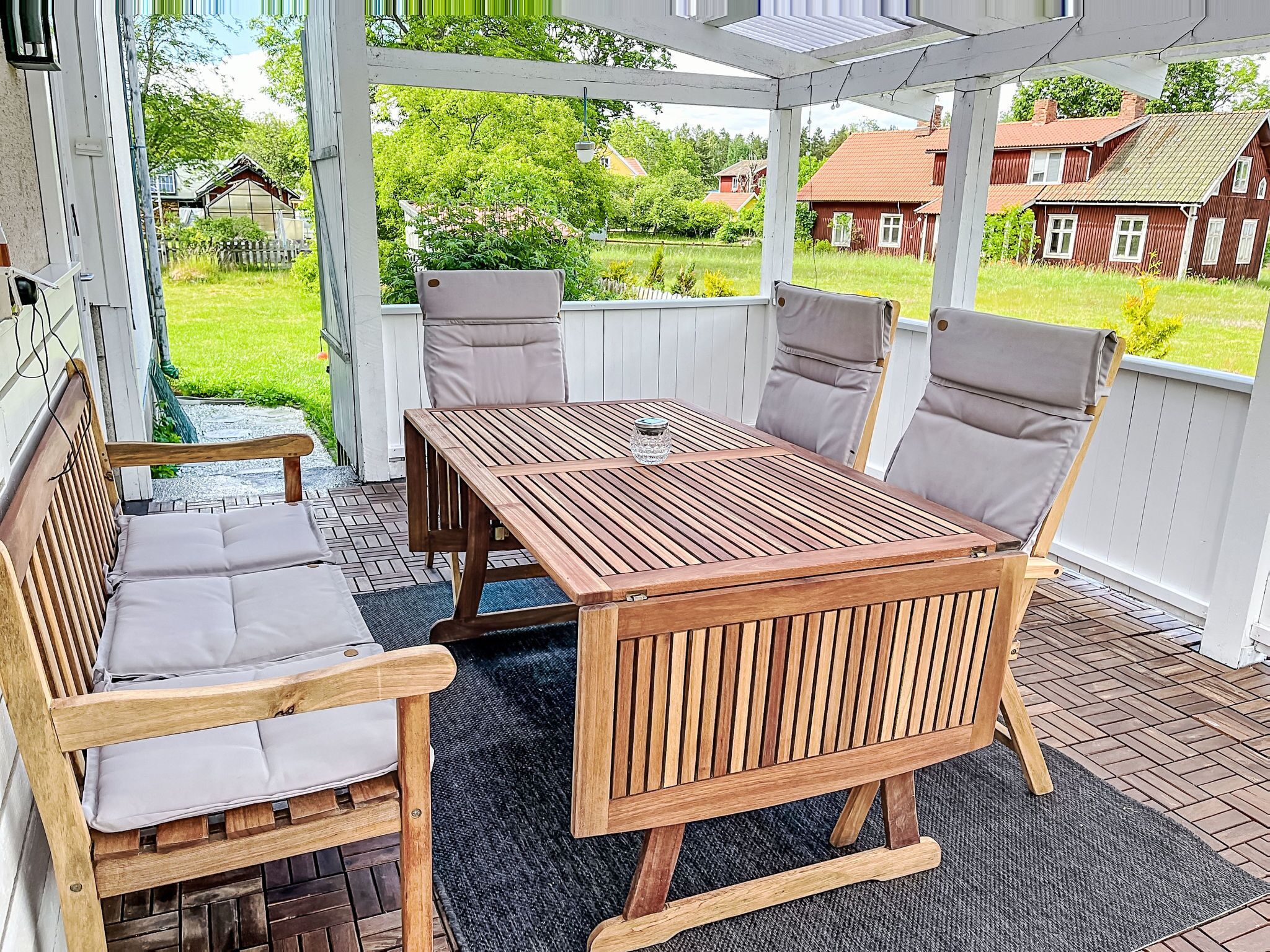 A wooden outdoor seating area with a table and chairs, a glass on the table, and a view of houses and greenery.