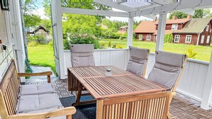 Ein hölzerner Außenbereich mit Sitzgelegenheit, Tisch und Stühlen, einem Glas auf dem Tisch sowie Blick auf Häuser und Grünflächen.