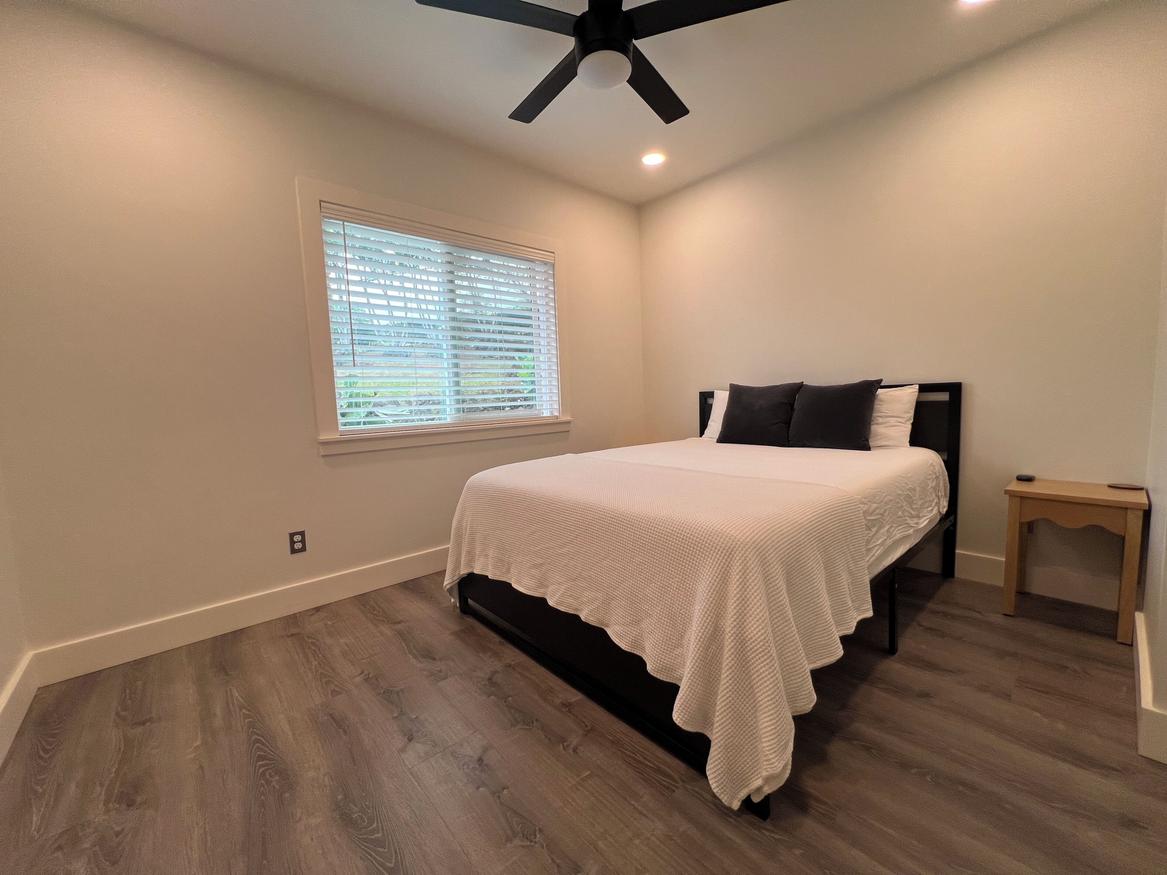 A bedroom with a bed, a window with blinds, a ceiling fan, and a small bedside table.