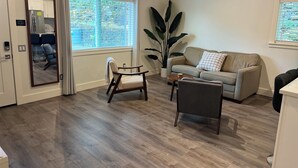 A living room with a sofa, armchairs, a coffee table, a large potted plant, and a sliding glass door leading to a kitchen.