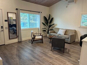 A living room with a sofa, armchairs, a coffee table, a large potted plant, and a sliding glass door leading to a kitchen.