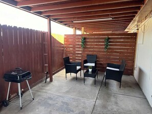 A covered patio with a wooden fence, a grill, and four wicker chairs with a small table.