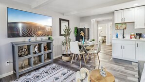 A modern living room with a large flat-screen TV, a dining area with a glass table, and a kitchen with white cabinets.