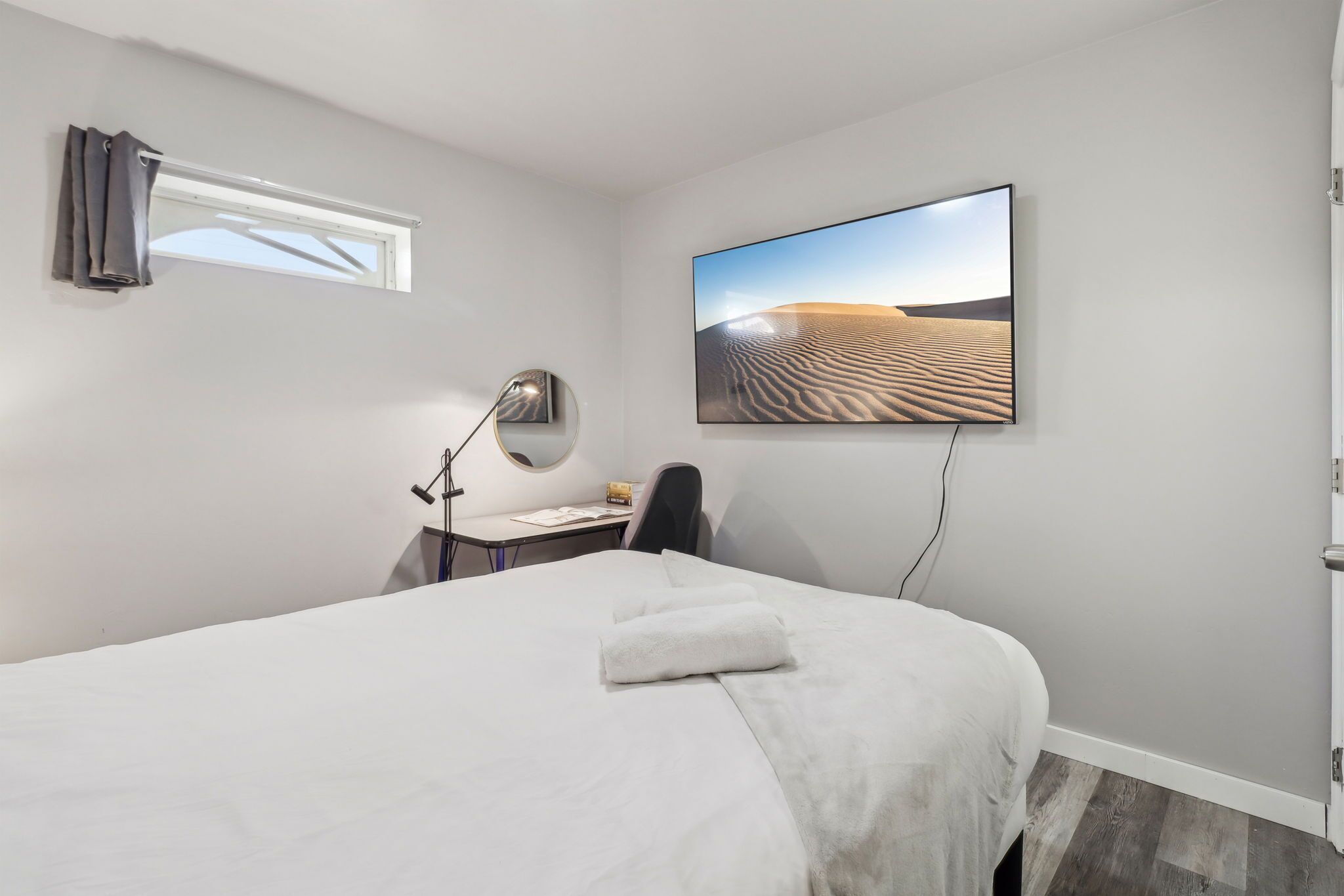 Une chambre d’hôtel avec un lit, un bureau, une chaise, une lampe de bureau, une fenêtre avec des rideaux et un tableau représentant un paysage désertique accroché au mur.
