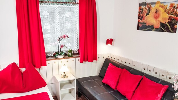 Quarto com cama, sofá, mesa de cabeceira e janela com cortinas.