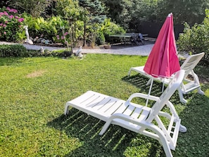 Ein Garten mit einer weißen Liege, einem roten Sonnenschirm und einem Tisch, umgeben von viel Grün.