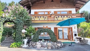 En toetages chalet med balkon, sten trapper der fører op til indgangen, og en blå parasol.