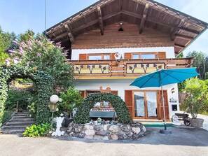 Eine zweistöckige Chalet mit Balkon, Steinstufen zum Eingang und einem blauen Sonnenschirm.