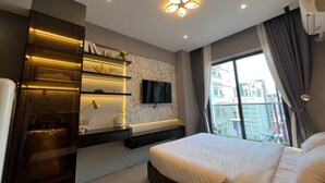 A modern bedroom with a large bed, built-in shelving, a flat-screen TV, and a city view.