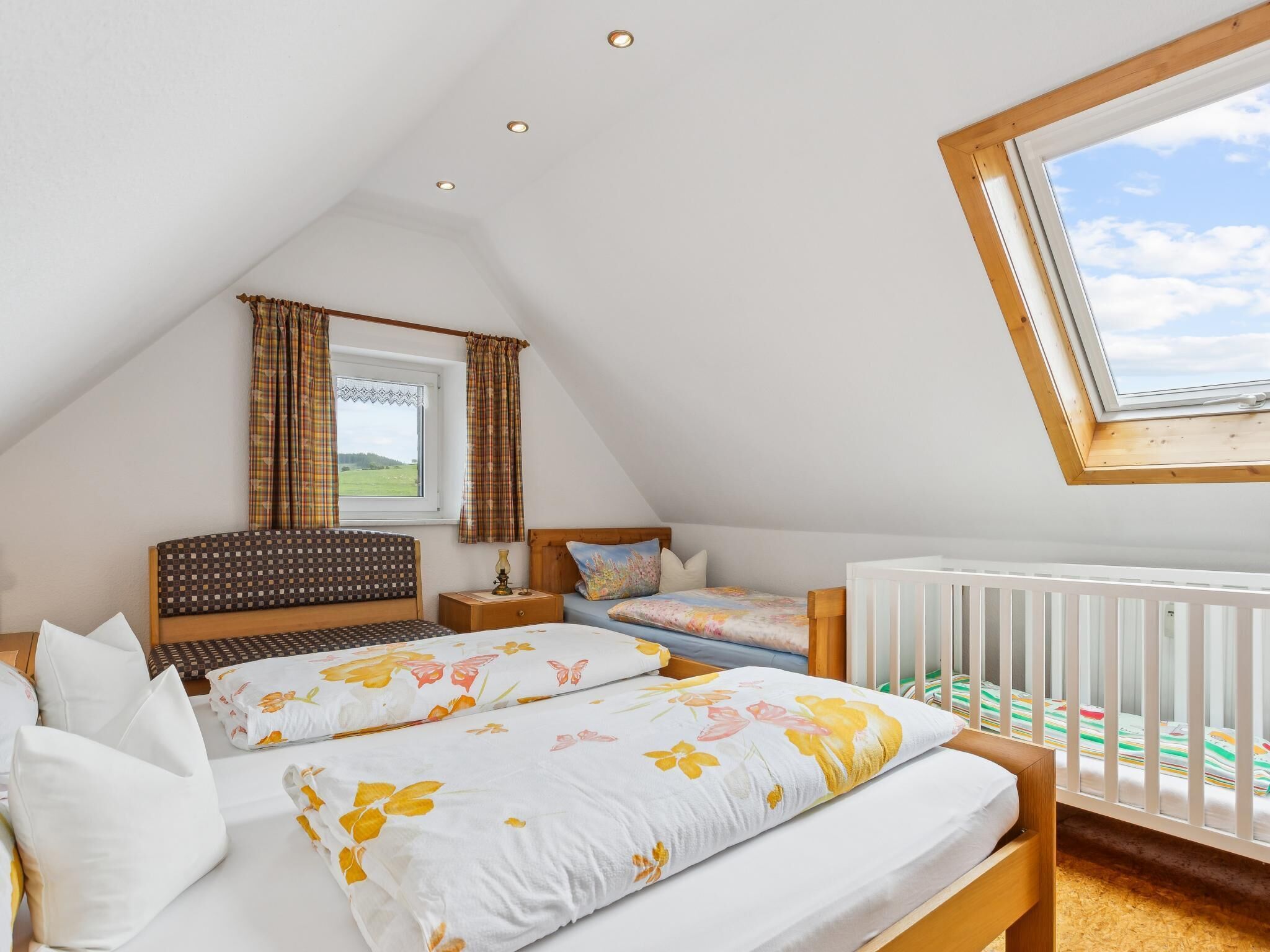 Une chambre avec un plafond en pente, une fenêtre avec des rideaux, un berceau et un lit avec une literie à motifs floraux.
