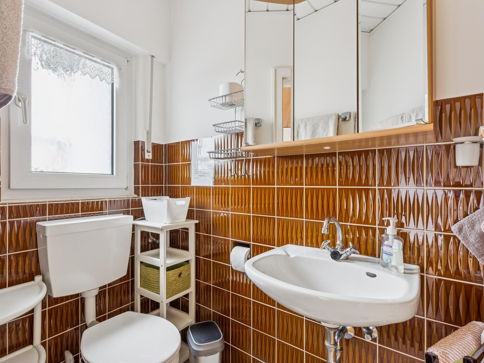 A bathroom with a white sink, toilet, and bidet, a window with lace curtains, and a mirror above the sink.