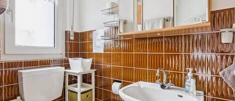 A bathroom with a white sink, toilet, and bidet, a window with lace curtains, and a mirror above the sink.