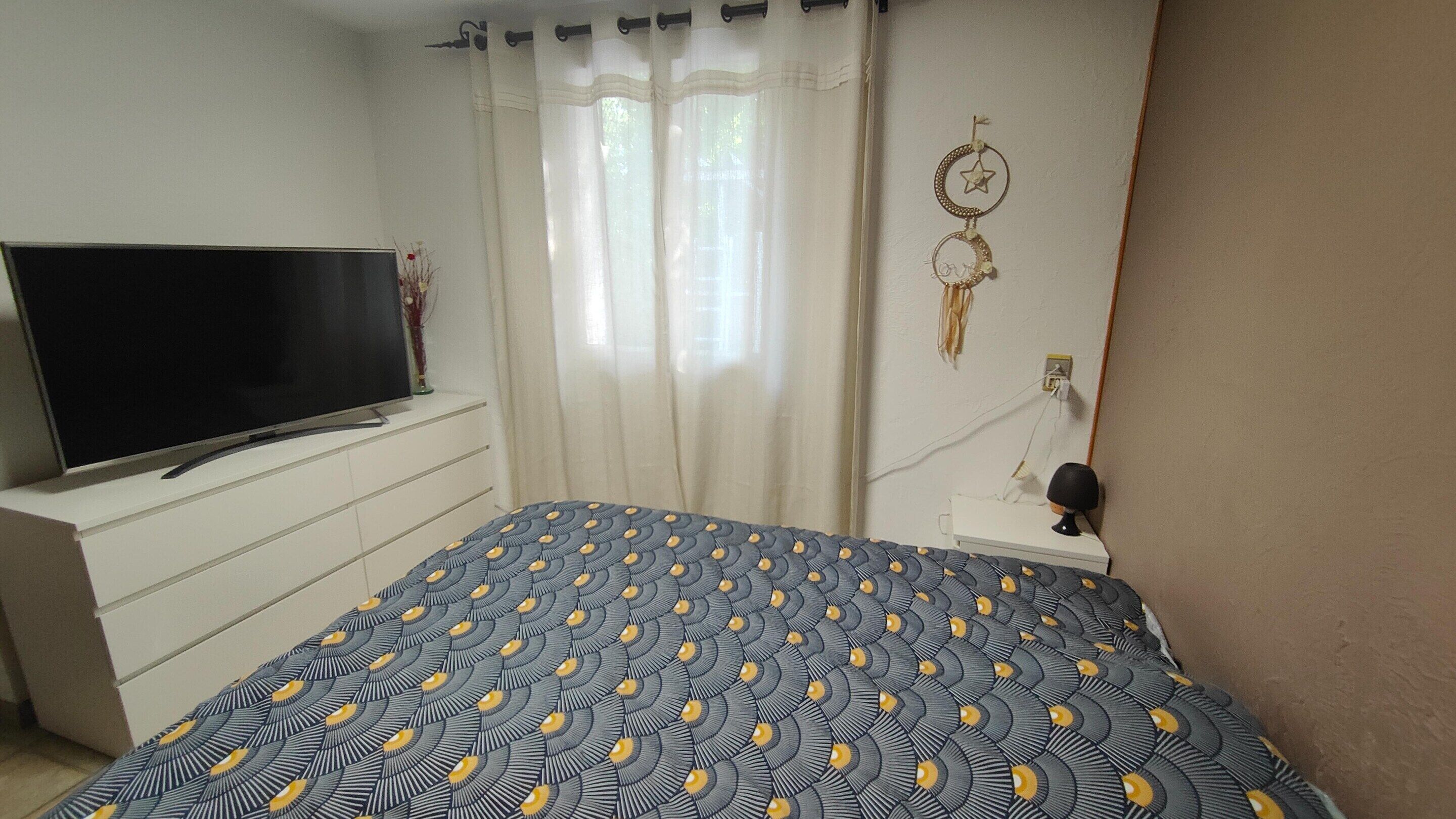 Un dormitorio con cama, televisor sobre una cómoda, lámpara de pared y ventana con cortinas.