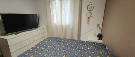 Un dormitorio con cama, televisor sobre una cómoda, lámpara de pared y ventana con cortinas.
