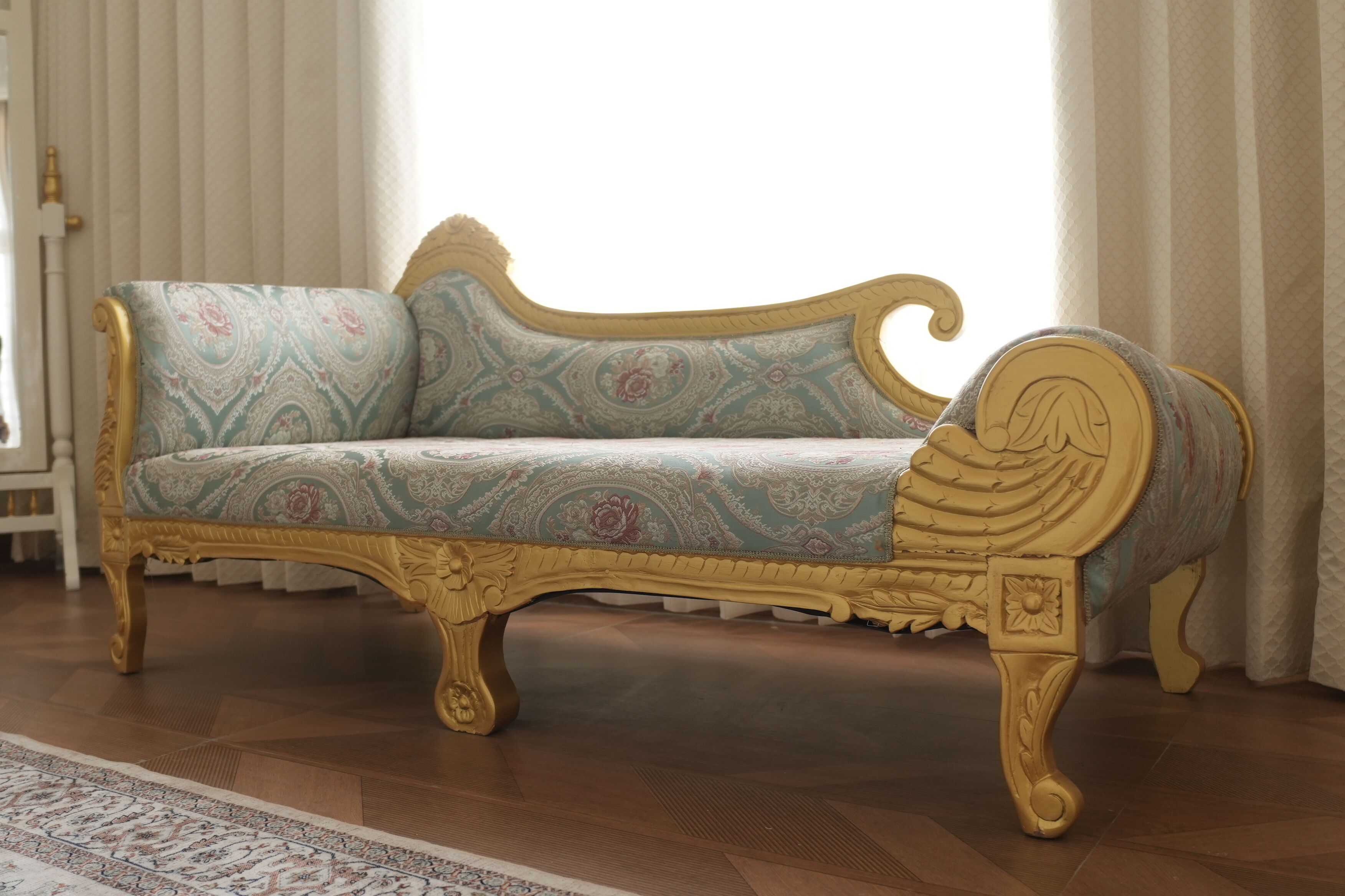 Un sofá antiguo con detalles dorados ornamentales y tapicería estampada en una habitación con suelo de madera y alfombra con diseño.