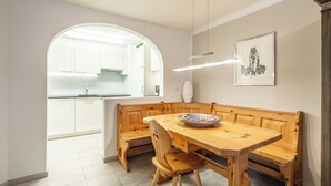A dining area with a wooden table and benches, a white kitchen in the background, and a framed artwork on the wall. - Runca 752 by Interhome (Flims)