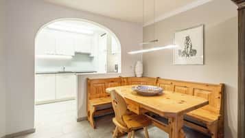 A dining area with a wooden table and benches, a white kitchen in the background, and a framed artwork on the wall.