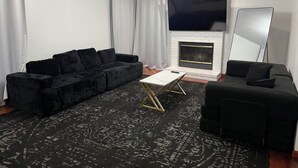 Un salon avec un canapé noir, une table basse blanche, une cheminée, un téléviseur et un grand miroir.