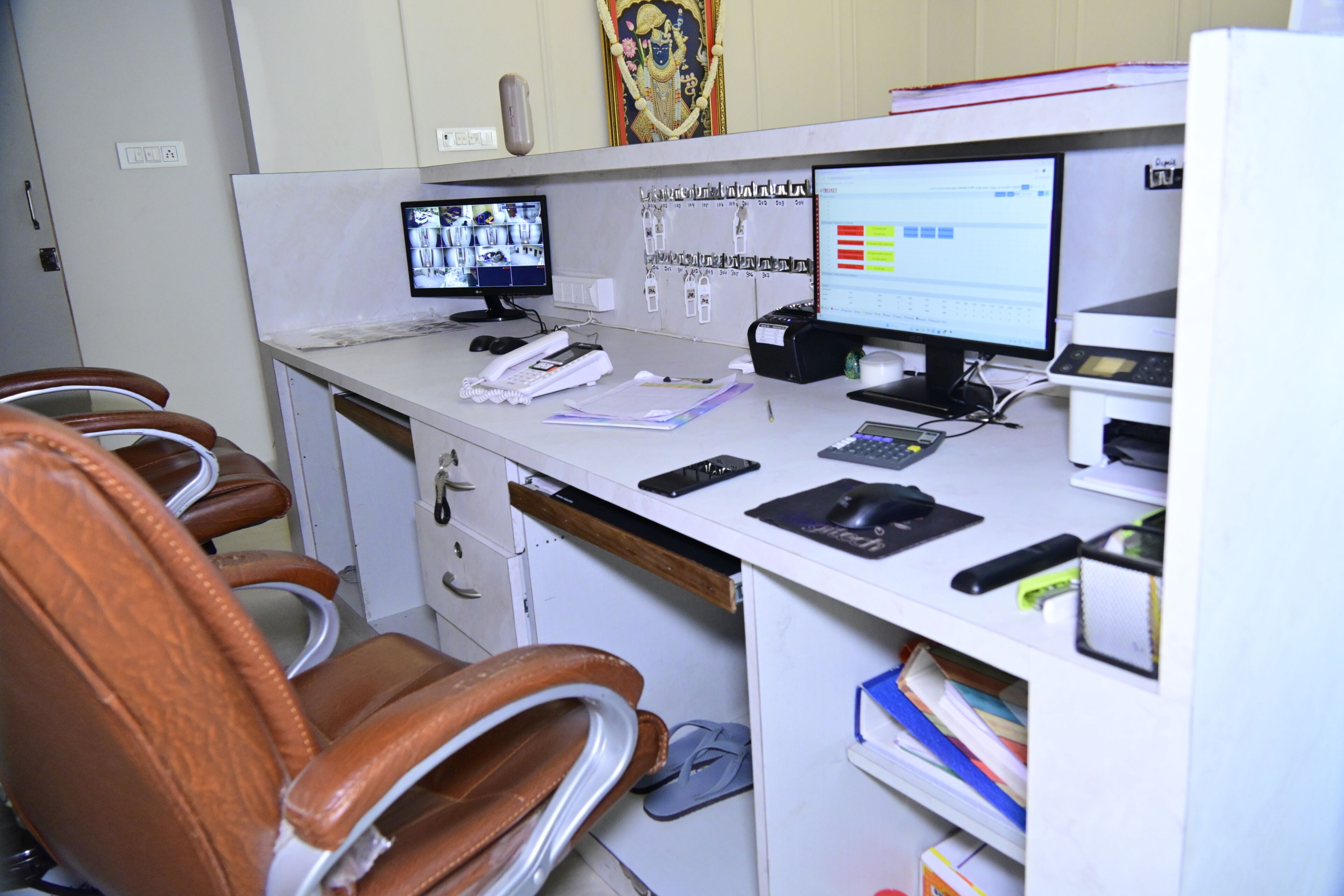 Um espaço de trabalho com dois monitores de computador, um telefone e um teclado. Há cadeiras de couro marrom e uma mesa branca.