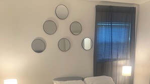 Une chambre à coucher comprenant un lit, deux tables de chevet, un miroir rond, une fenêtre avec des stores et une lampe sur chaque table.