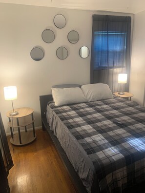 A bedroom with a bed, two bedside tables, a round mirror, a window with blinds, and a lamp on each table.