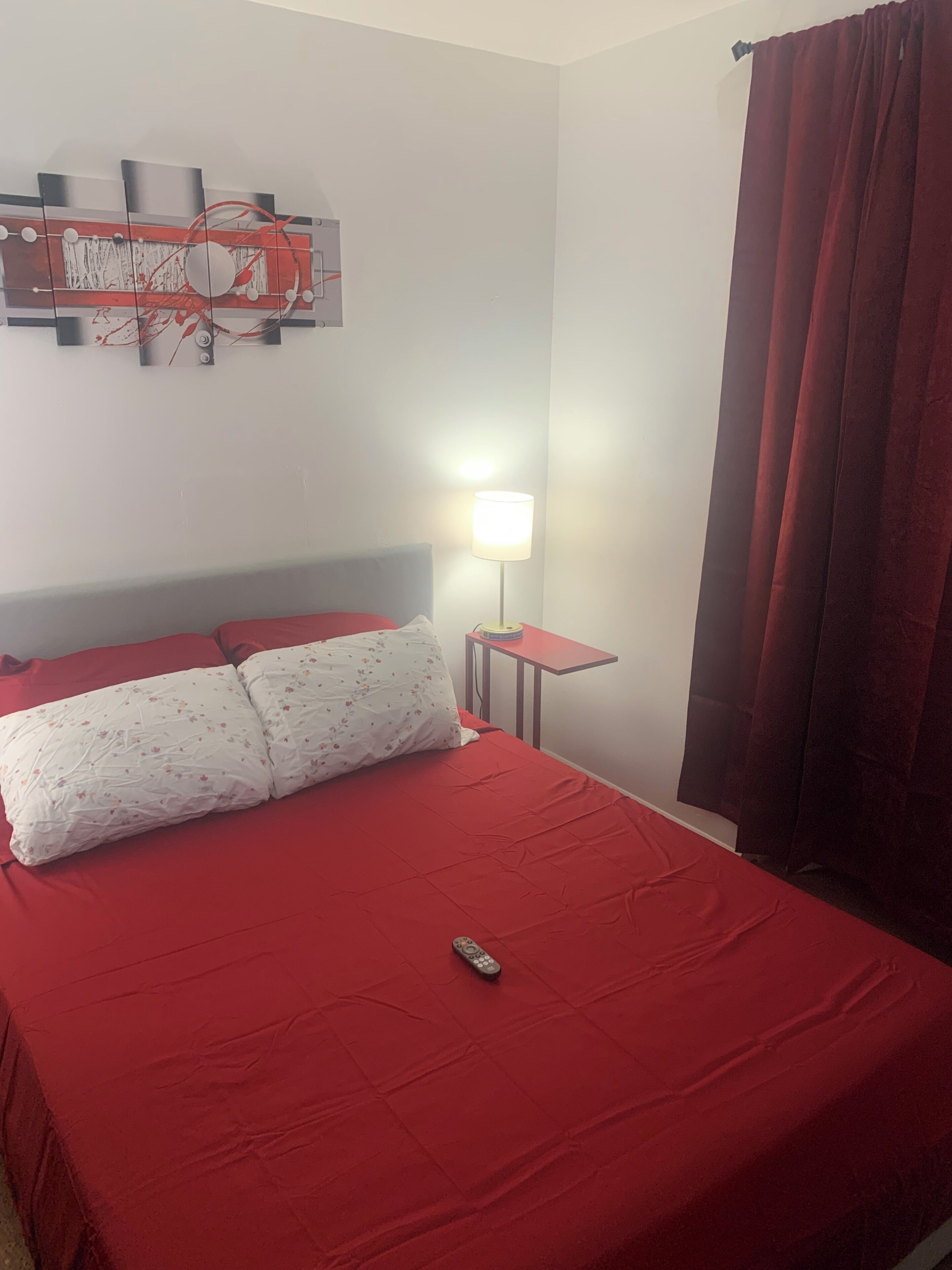 A bed with red bedding, a remote control, a lamp, and a wall decoration.
