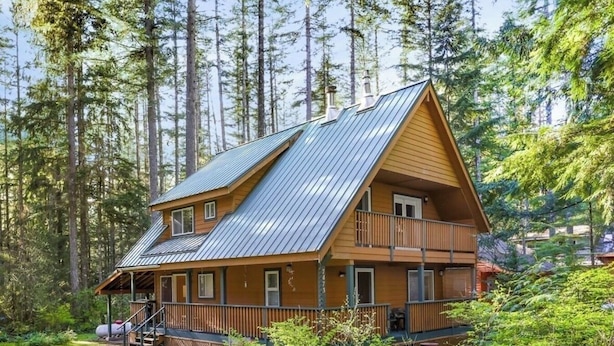Une cabane en rondins à deux étages, surmontée d’un toit en métal, entourée d’une forêt dense.