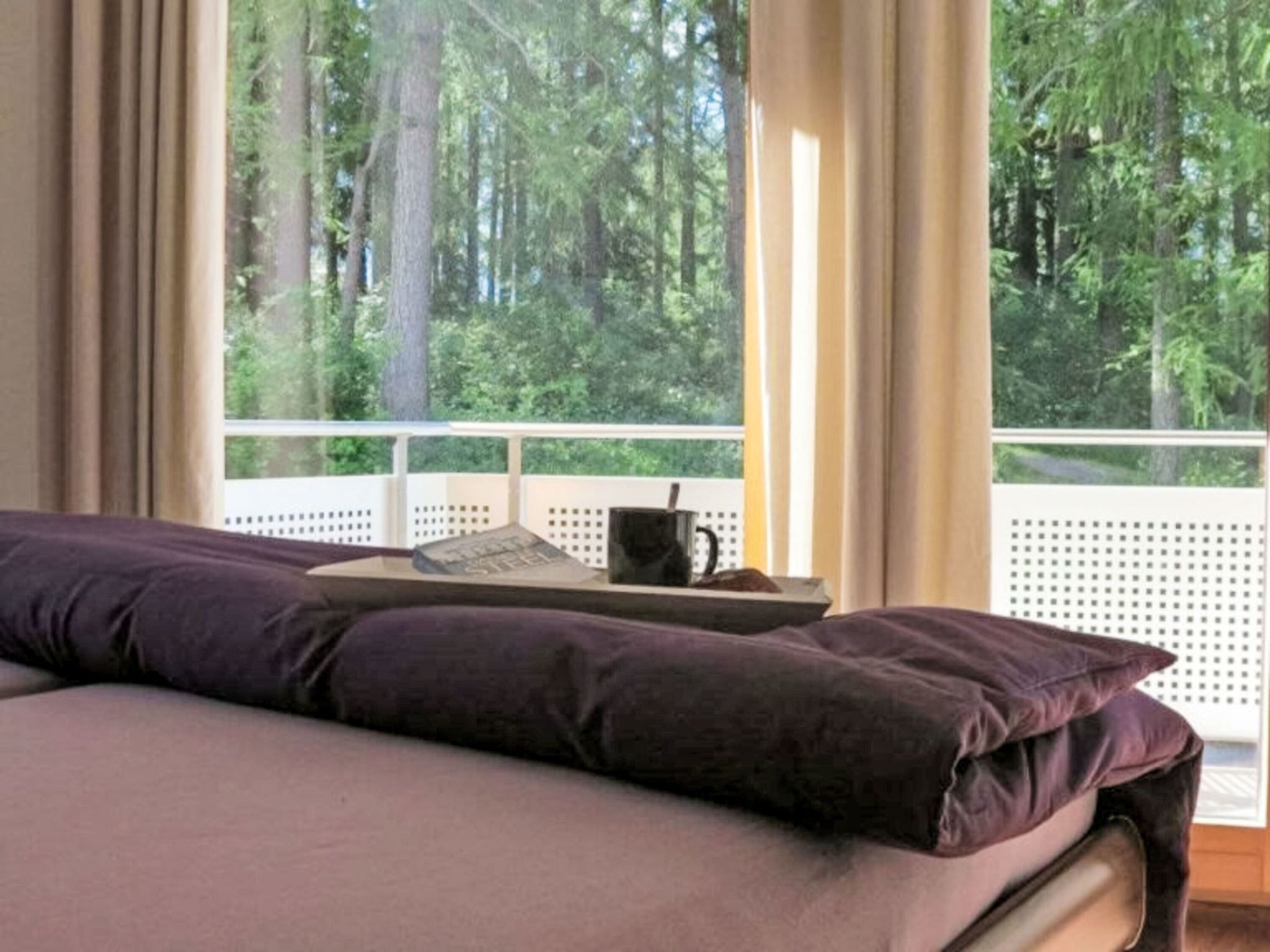 Ein Schlafzimmer mit einem Bett, einem Fenster mit Blick auf Bäume, einer Tasse und einem Buch auf einem Nachttisch.