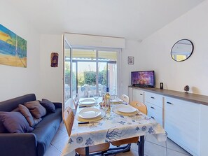 A dining area with a table set for four, a sofa, a TV, and a door leading to an outdoor patio. - Le Tropique by Interhome (Saint Cyprien)
