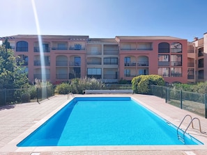 Une piscine avec une terrasse carrelée, entourée d’une clôture, et un immeuble à plusieurs étages avec des balcons en arrière-plan.