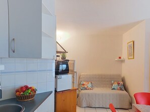 A compact living space with a kitchenette, a bed with floral pillows, and a red chair. - Les Goelettes by Interhome (Saint Cyprien)