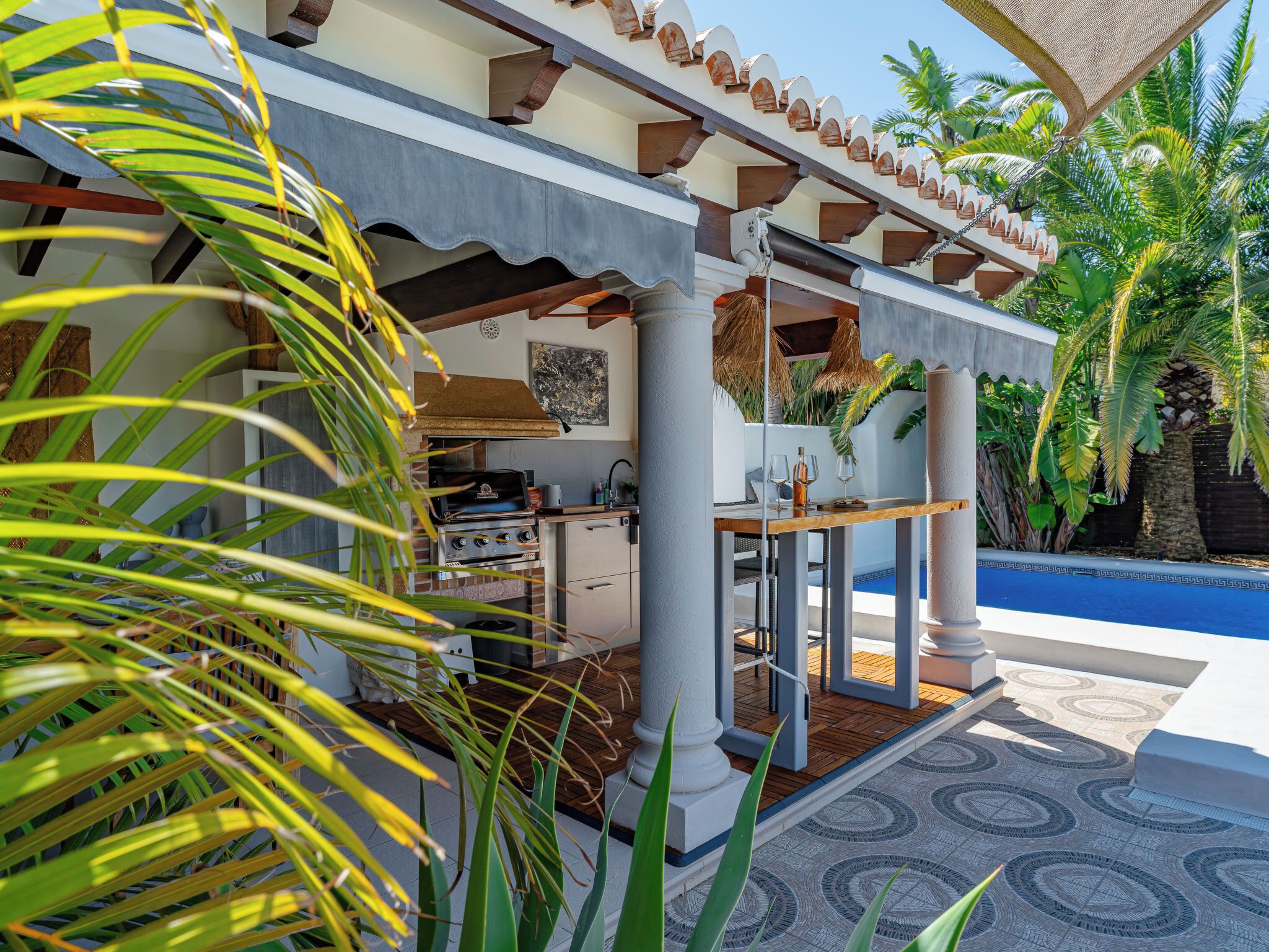 A covered outdoor kitchen with a grill, a dining table, and a view of a pool.