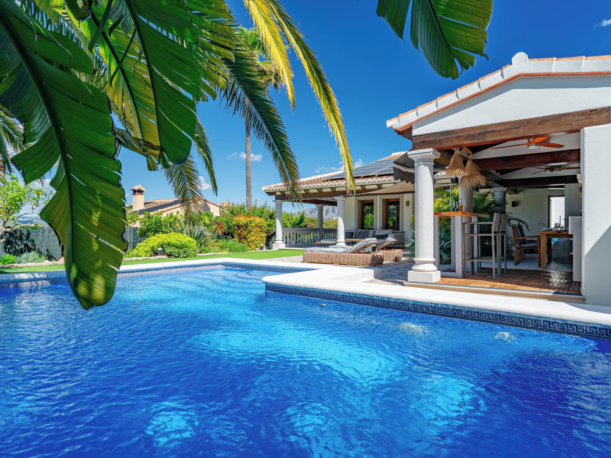 A swimming pool with a pergola and lounge chairs, surrounded by lush greenery and a clear blue sky.