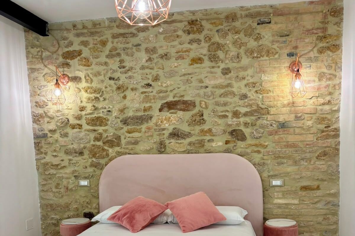 A bedroom with a stone wall, a bed with pink bedding, two bedside tables, and a hanging light fixture.