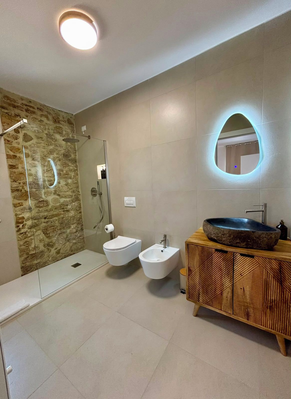 Un baño moderno con pared de piedra, cabina de ducha de vidrio, un bidé, lavamanos con lavabo de piedra y tocador de madera.