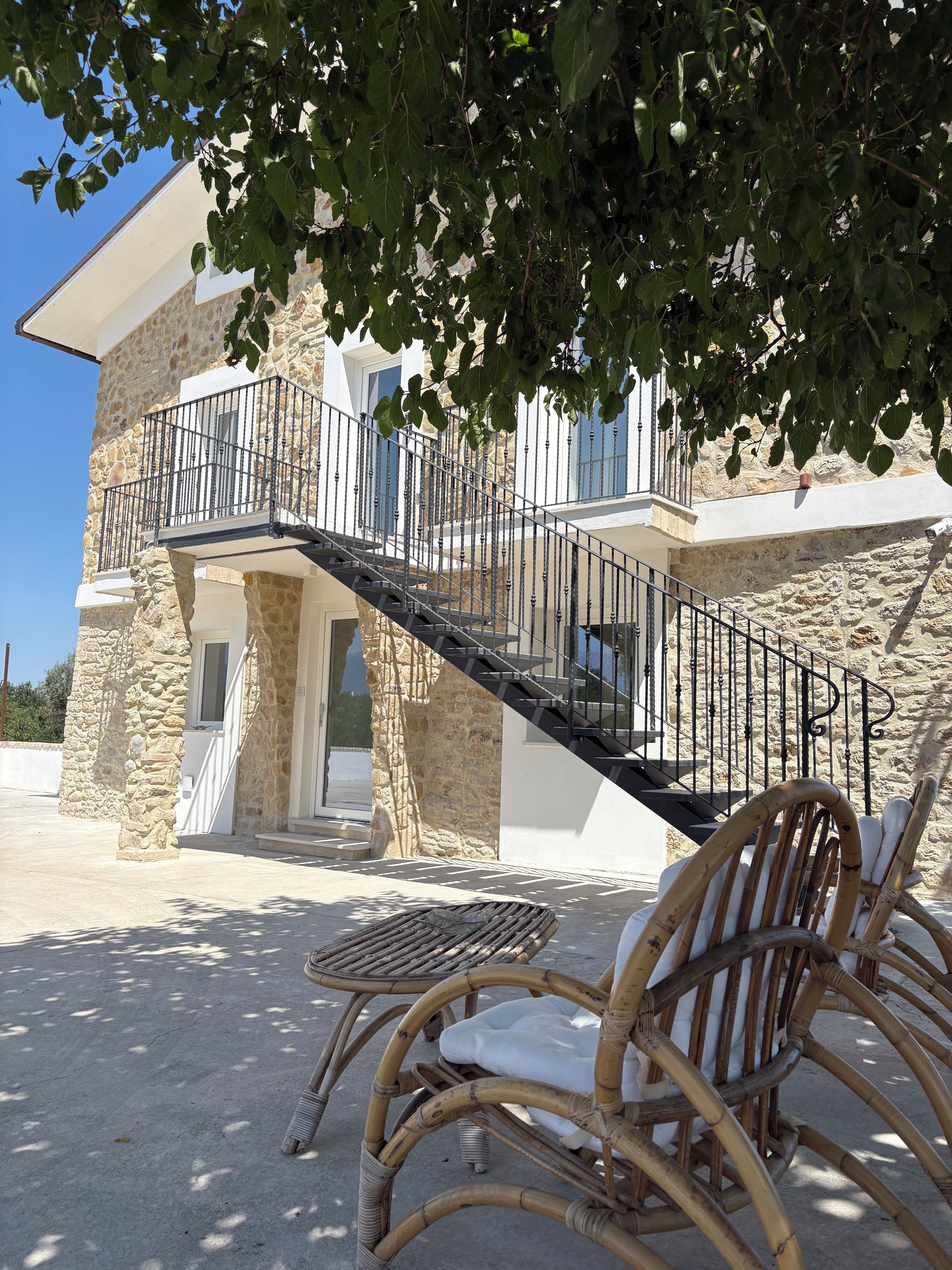 A stone building with a balcony and a staircase leading to a second floor. There is a wicker chair and a table in the foreground.