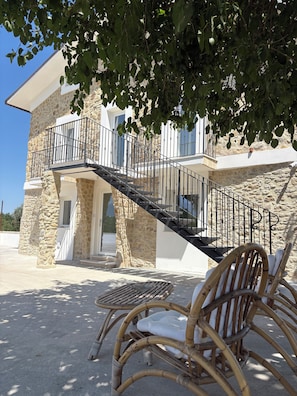 A stone building with a balcony and a staircase leading to a second floor. There is a wicker chair and a table in the foreground.