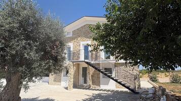 A stone building with a staircase leading to a balcony, surrounded by olive trees and a clear sky.