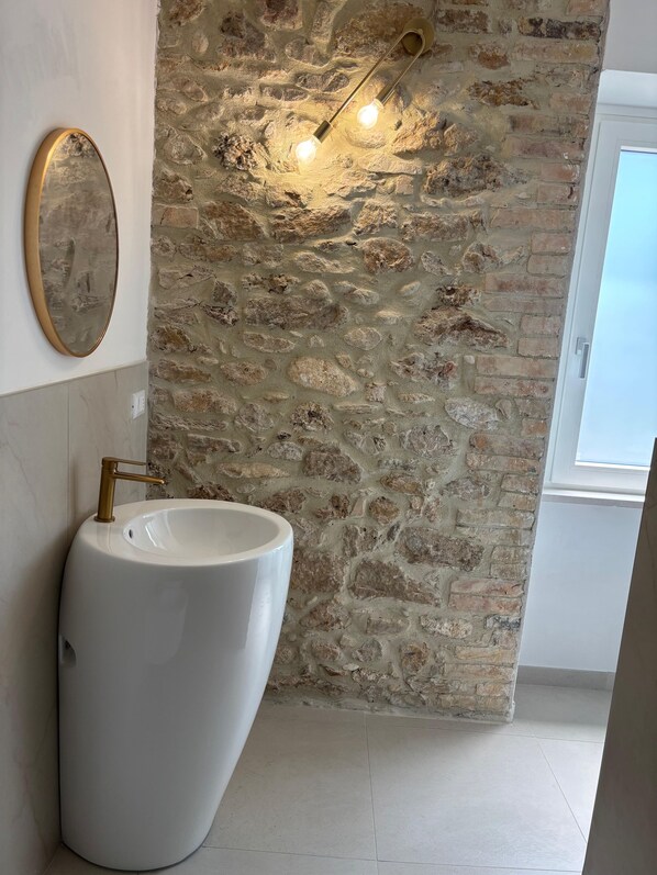 A bathroom with a white sink, gold faucet, and a stone wall. There are two wall-mounted lights and a window.