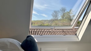 Un dormitorio con tragaluz, una cama con almohadas y vistas a un tejado de tejas y árboles.