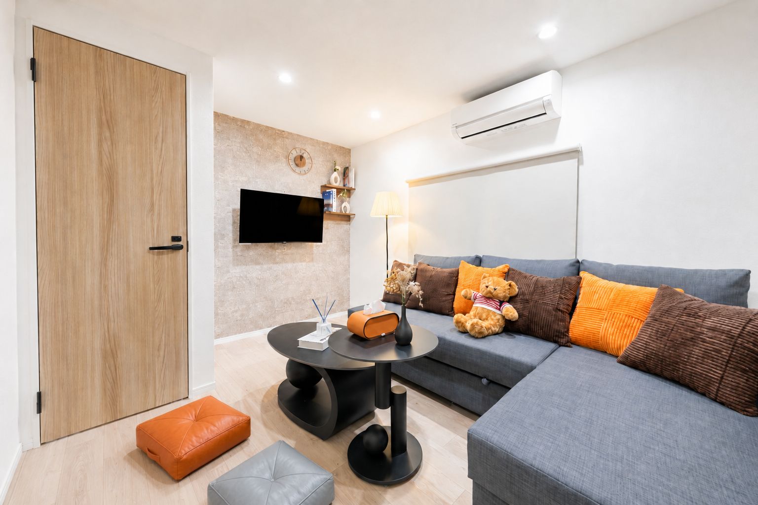 A modern living room with a grey sofa, orange and brown cushions, a round black table, and a television mounted on the wall.
