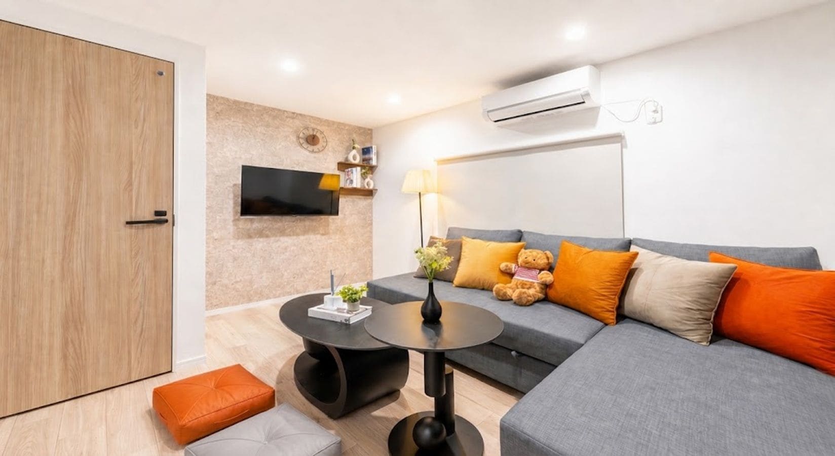 A modern living room with a grey sofa, orange cushions, a round coffee table, and a flat-screen TV mounted on the wall.