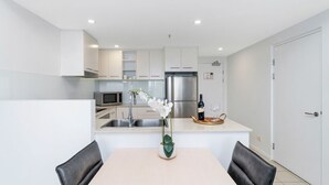 Eine moderne Küche mit weißen Schränken, einem Edelstahlkühlschrank, einer Mikrowelle und einem Spülbecken. Es gibt einen Esstisch mit zwei Stühlen und eine Vase mit weißen Blumen.