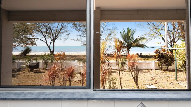 Vista attraverso una finestra su una proprietà vicino alla spiaggia, con barbecue, palme e una recinzione.