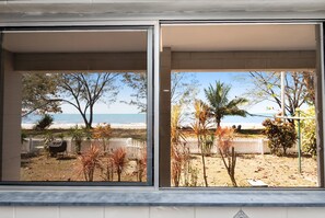 En vy genom ett fönster i en villa vid stranden, med en grillplats, palmer och ett staket.