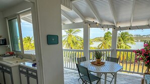 Een keuken met een spoelbak en kasten, een eethoek met een tafel en stoelen, en een balkon met uitzicht op palmbomen en water.
