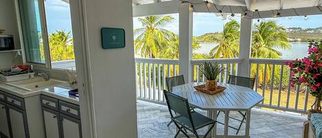 A kitchen with a sink and cabinets, a dining area with a table and chairs, and a balcony with a view of palm trees and water.